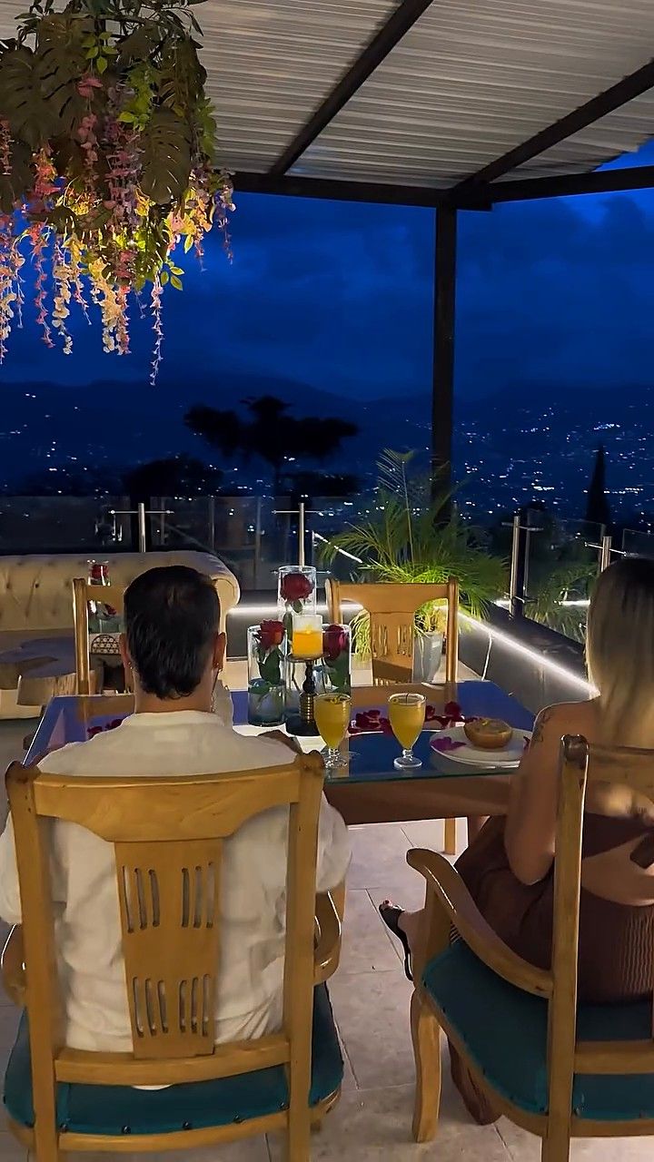 Mesa servida para dos en la terraza de Garden SPA, con jugos y pétalos, y la ciudad iluminada al fondo al anochecer