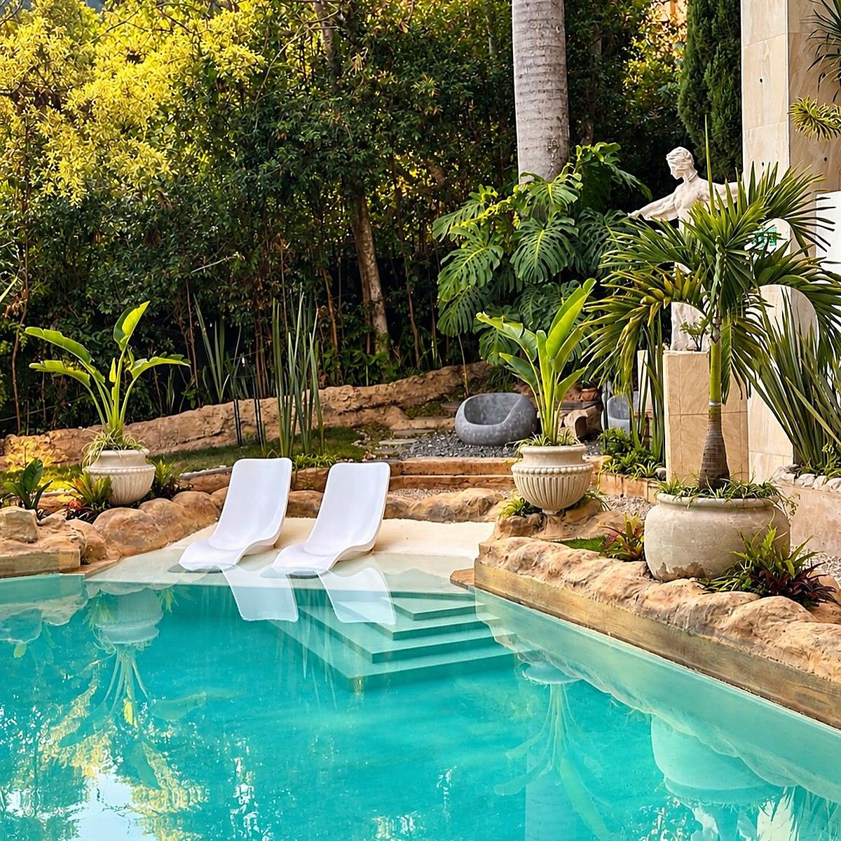Piscina de agua turquesa de Garden SPA rodeada de rocas, palmas y jardín, con dos asoleadoras blancas dentro del agua
